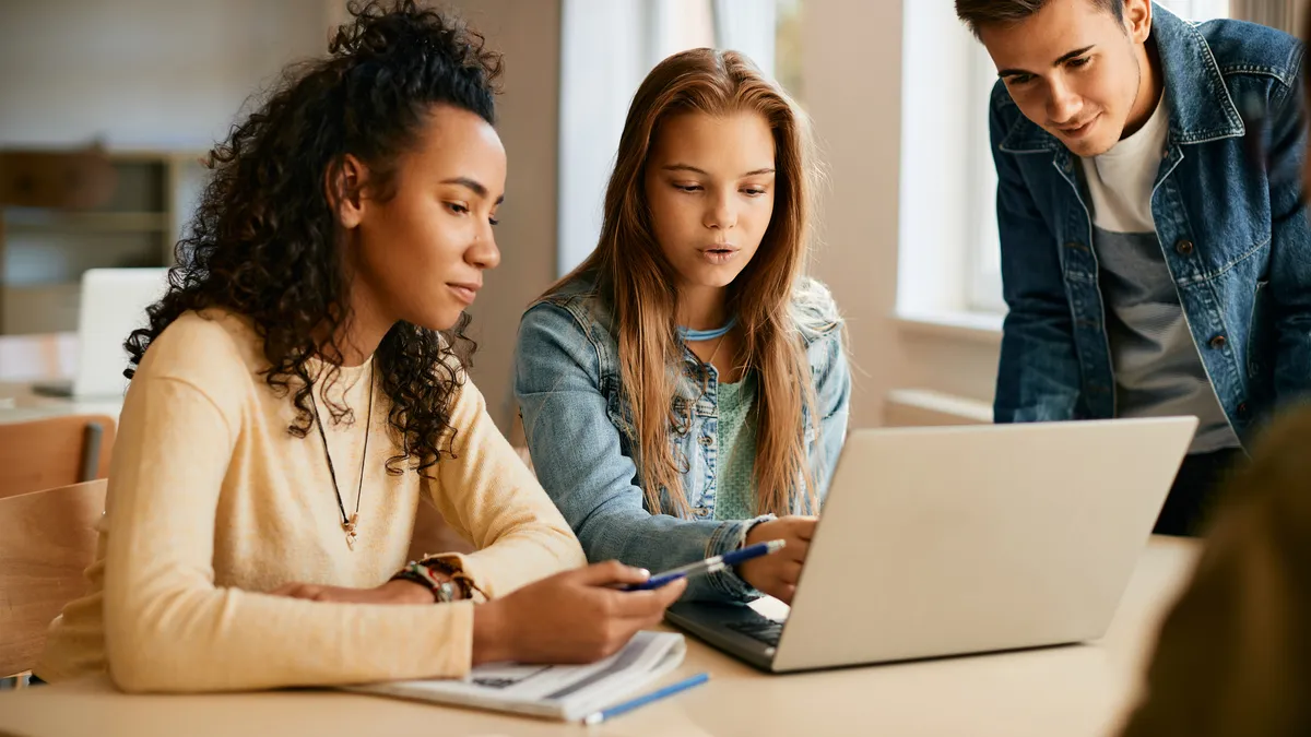 Two female students are seated looking at a computer as a male peer leans over the table to look at their work.