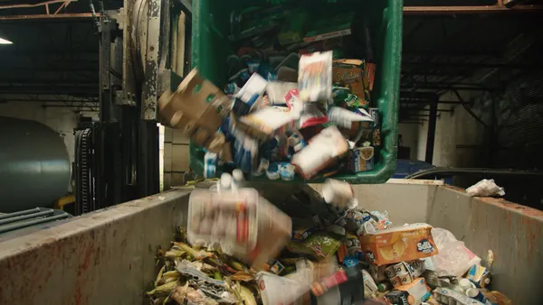 A large bin full of packaged, uneaten food is tipped into a dumpster by a machine inside a facility.