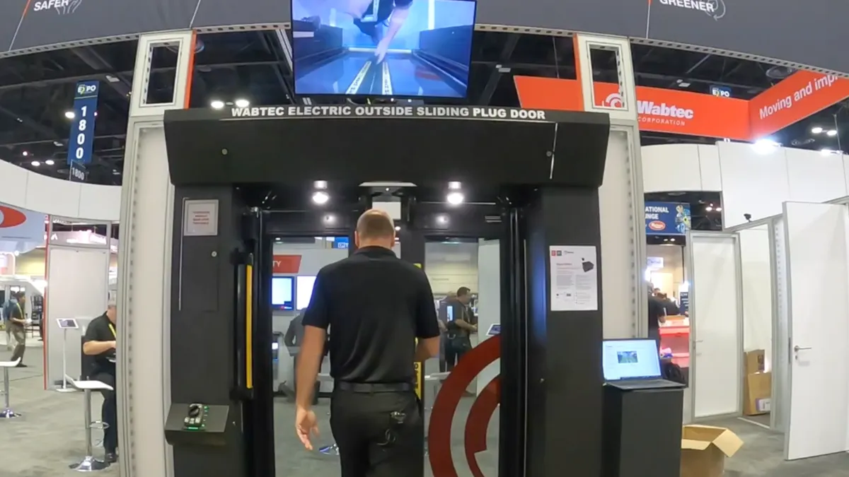 A man seen from behind, dressed in black, walks though a mockup of a bus door in an exhibit hall.