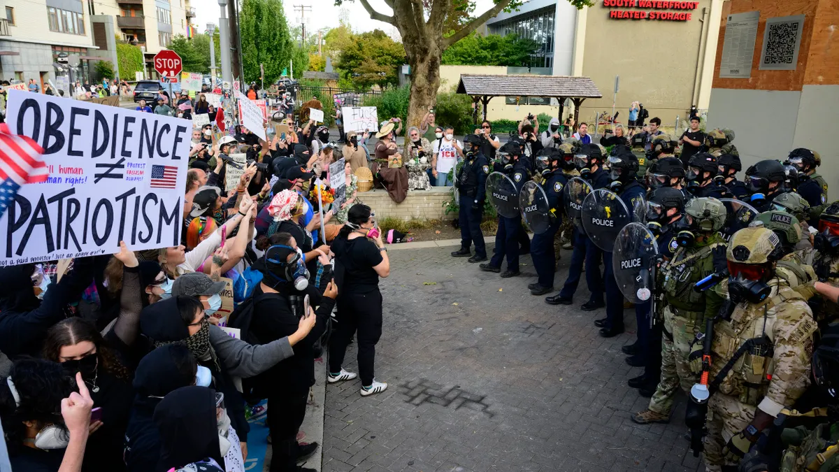 A group of people holding signs faces a group of officers in uniforms holding shielfs.