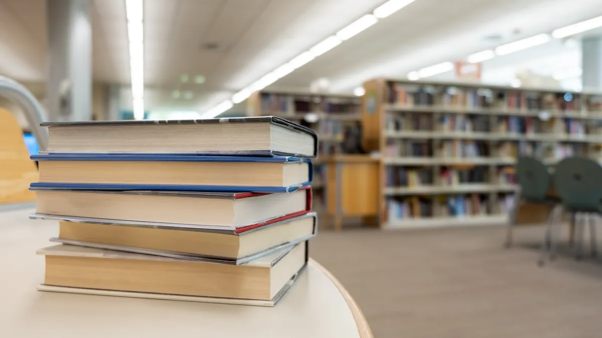 A stack of books sits on a table in the foreground. In the background are rows of shelves of books in a room