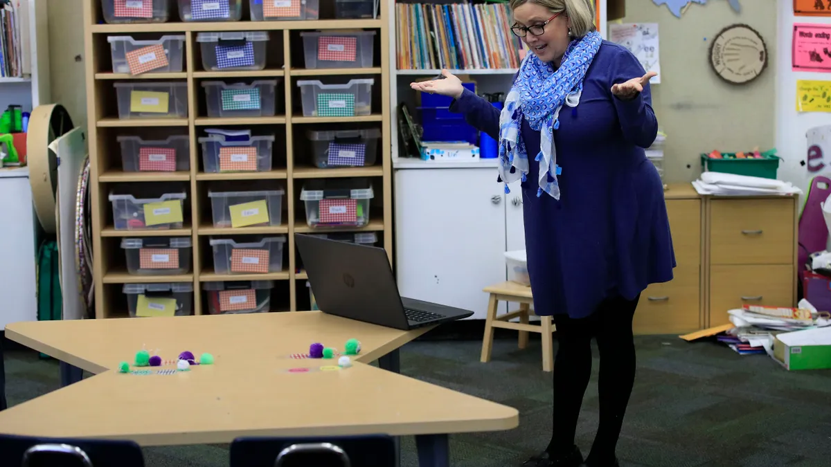 An adult stands in a classroom alone. There is a laptop on a desk in front of the person and the person is gesturing with their arms.