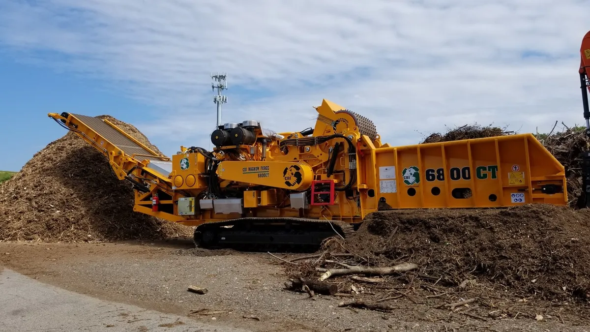 A large machine with a conveyor system and rotating cylindrical grinder sits outdoors between two piles of organic material.