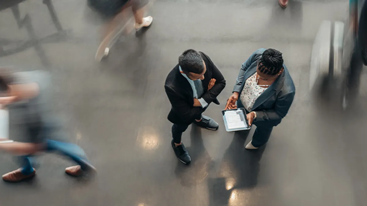 Two business people standing in the lobby of an office looking at a tablet while people are walking past in a blur