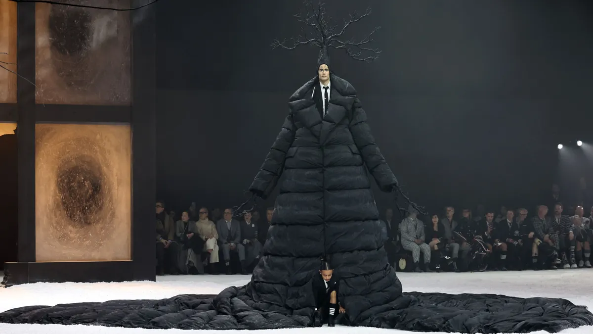 A child emerges from underneath an enormous black coat worn by a model at a fashion show.