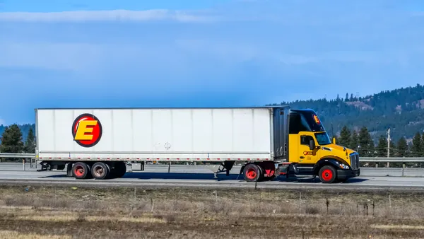 An Estes Express tractor-trailer travels on an interstate during the day with trees in the background.