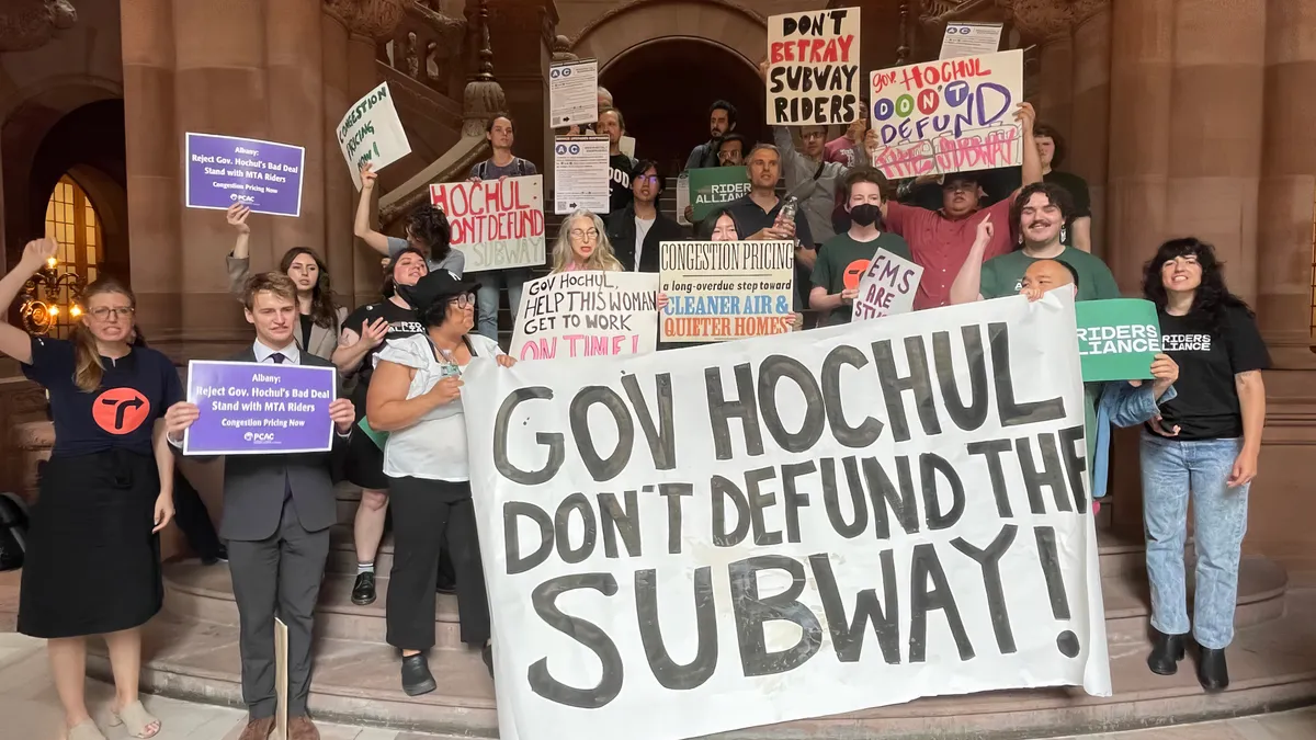 People holding signs in an ornate building. The largest sign reads, "Gov Hochul don't defund the subway."