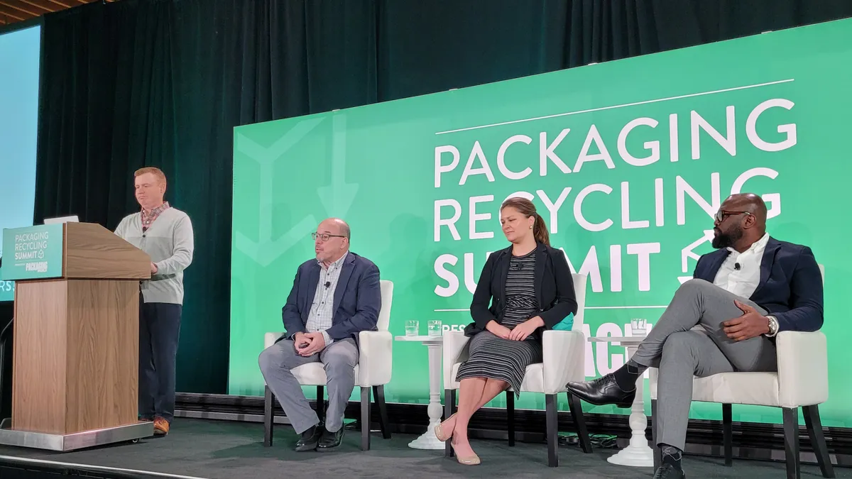 Three people sit in chairs on a stage and one person stands at a podium, with a green background behind all of them that says Packaging Recycling Summit.