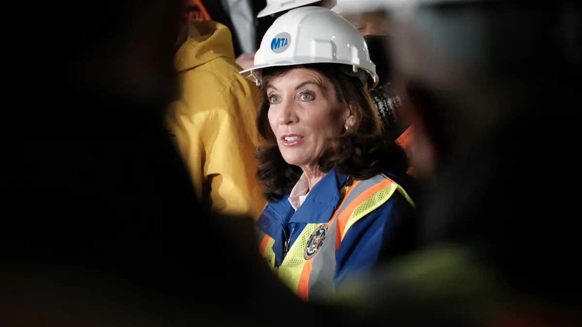 A woman wearing a white hard hat labeled "MTA" in a dark tunnel.