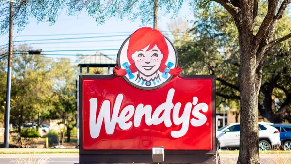 A red sign with white lettering that says Wendy's below a logo of a girl with red hair and blue hair ties sits in a parking lot under a tree.