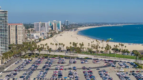 A parking lot full of cars and buildings line a sandy beach and blue water.