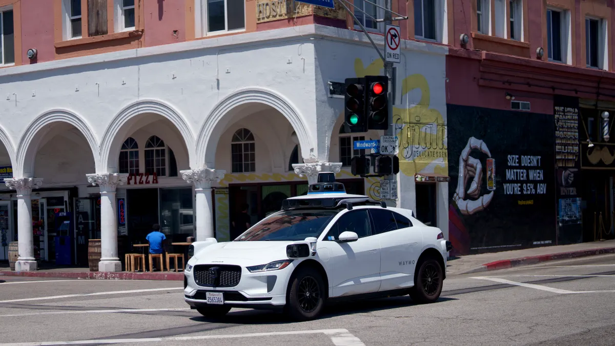 A white car with roof and side mounted sensors drives through an intersetion by a red building with a white facade and sign reading "pizza."