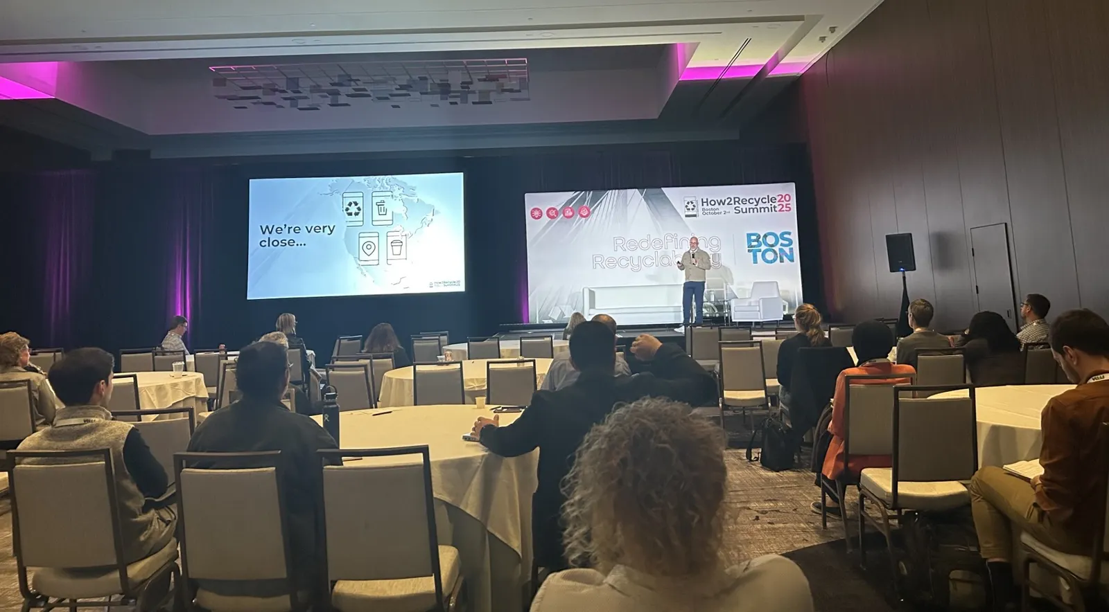 A person stands on stage in front of a room of people, next to a slide that reads "We're very close"