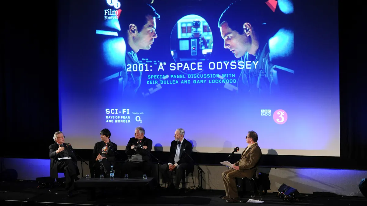 Four panelists and a moderator sit on a stage with an image showing two astronauts from 2001: A Space Odyssey.