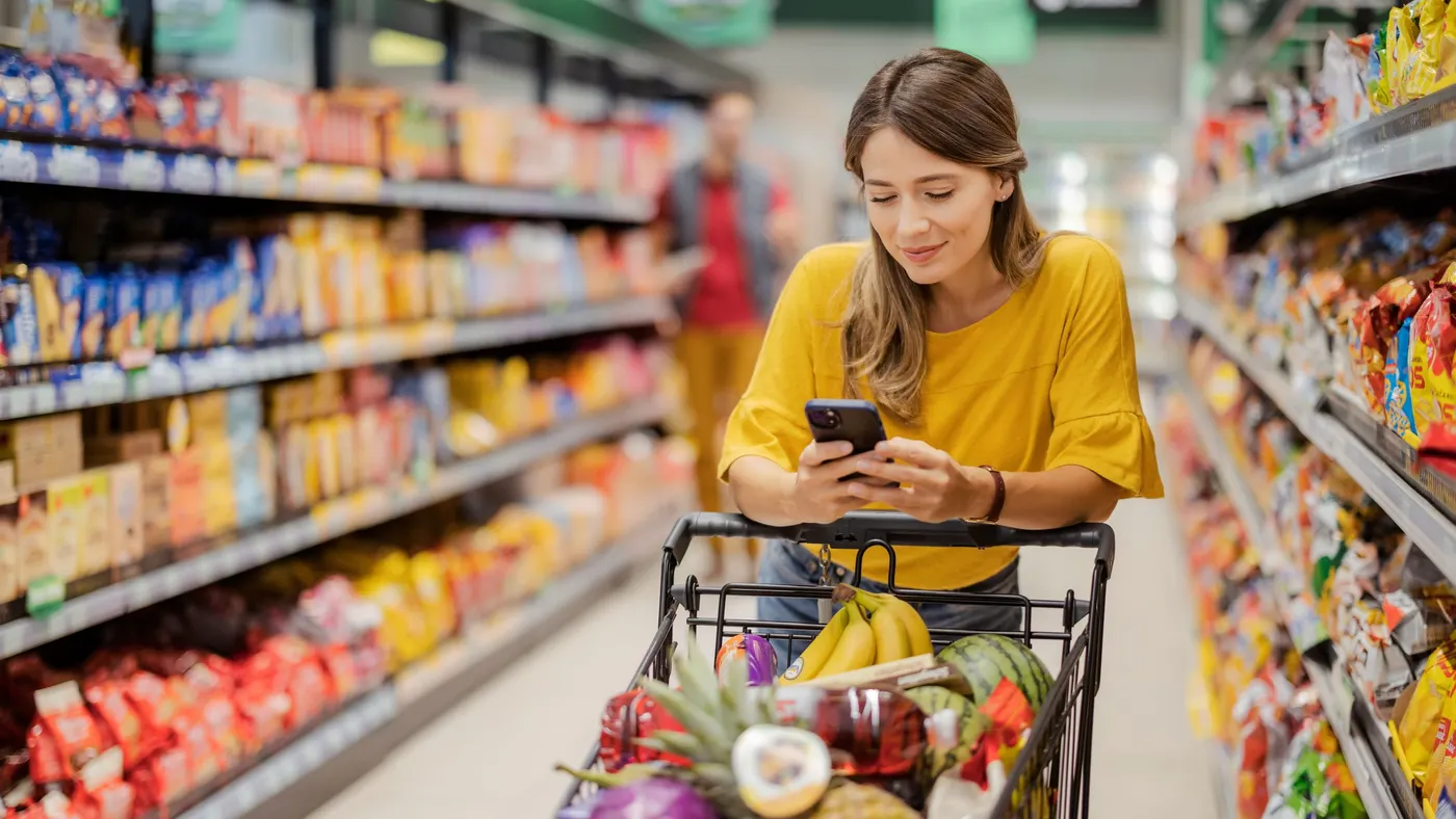 A person is in a grocery store pushing a cart filled with produce while they look at their phone.