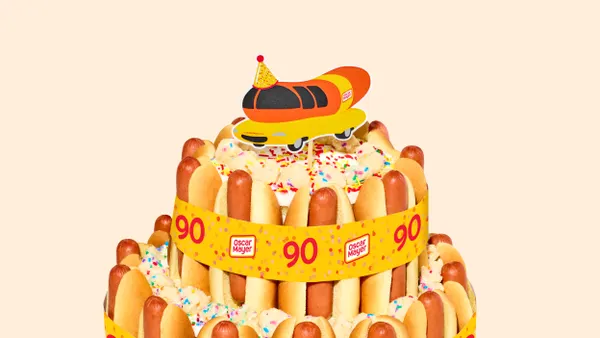 A two-tiered cake covered in hot dogs and topped with a Wienermobile cake topper.