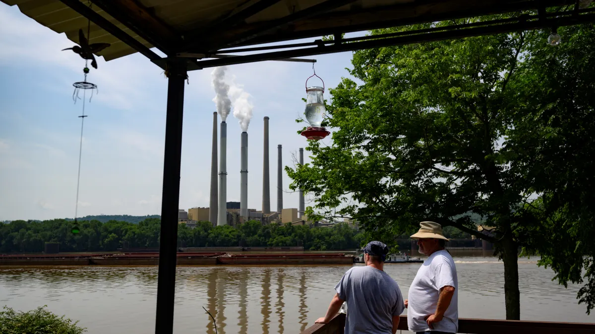 Two men stand near a river with power plant smokestacks in the background.