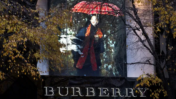 A sign featuring a person holding up a plaid umbrella can be seen through trees. Underneath the sign is the word 'Burberry" written in capital letters.