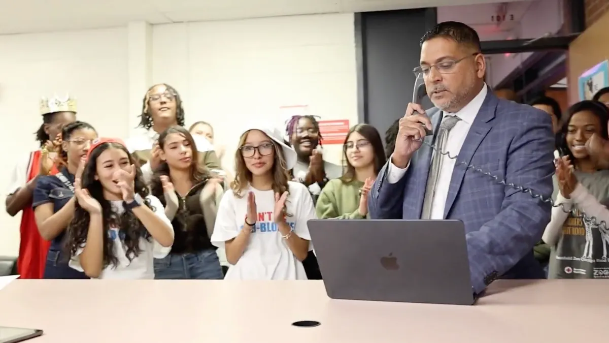 Superintendent Krish Mohip holds a phone while flanked by students in a school building office.