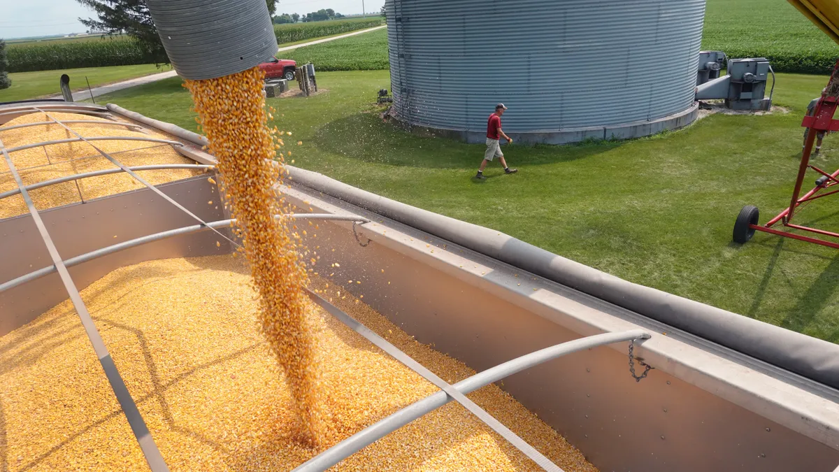 corn is unloaded into a truck as a person walks near a grain elevator in the background