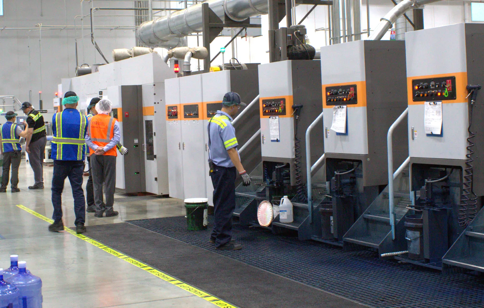 Numerous workers wearing protective gear stand near heavy machinery in an industrial facility.