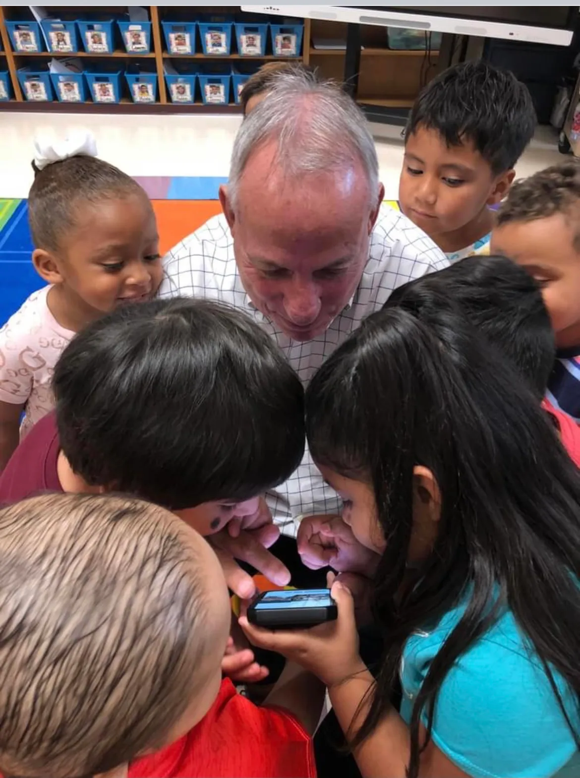 Adult looks down at a cellphone in hand while surrounded by young children also looking down at the phone. They are in a classroom with a bookcase.