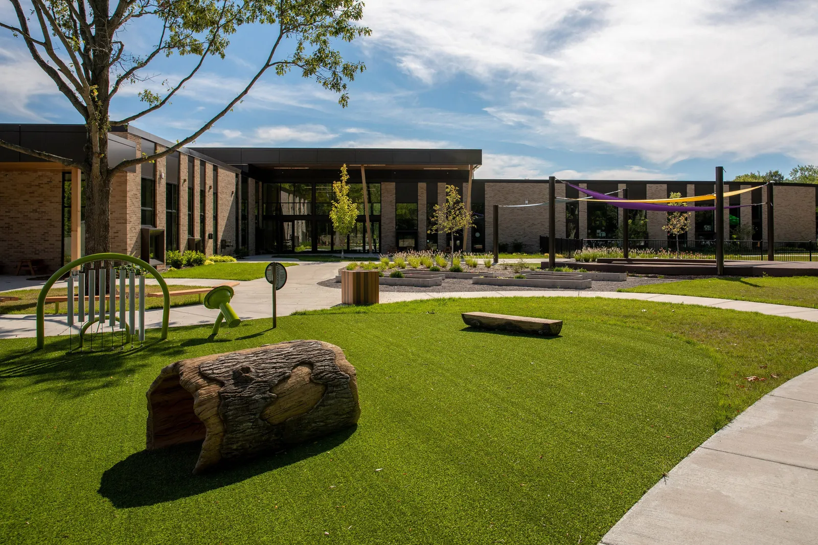 An outdoor play area is shown with play equipment and green turf surrounded by sidewalks. A brick and glass building is in the background.