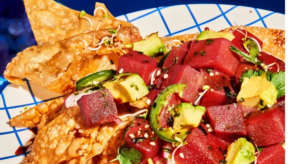 Ceviche with plant-based tuna chunks from Current Foods, avocado, pepper slices, herbs and sesame seeds on tortilla chips on a checkered plate with a blue background.