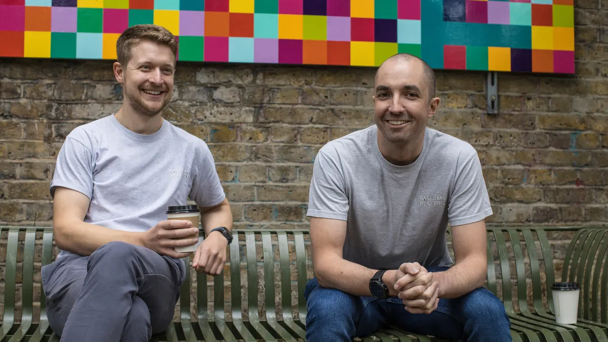 Two men sit on a bench, one of whom is holding a coffee cup.