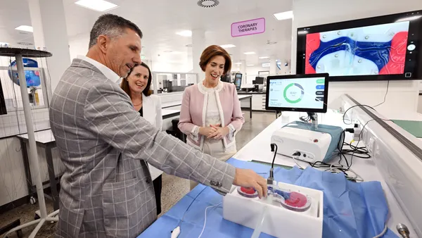 A person in a suit touches a square device with a tube while two other people watch.