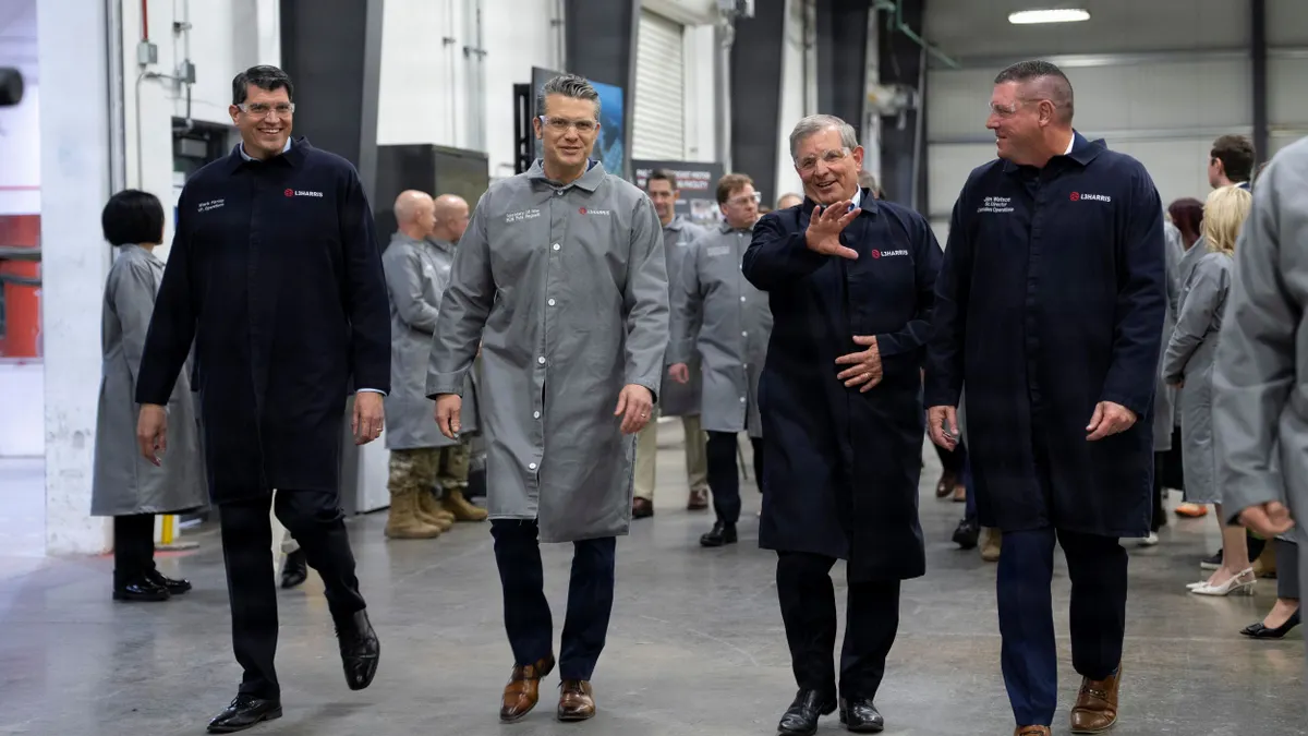 Four people wearing protective eyewear and labcoats walking side by side in a horizontal line inside a factory.