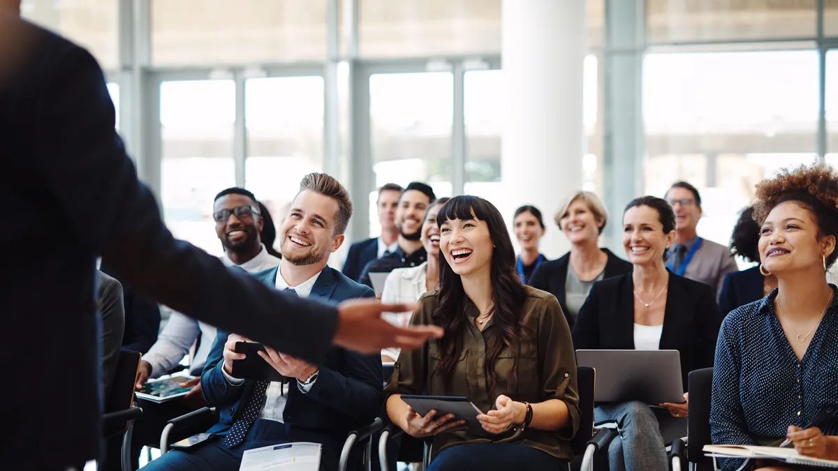 A diverse audience of smiling and laughing businesspeople are shown sitting in on a conference keynote. The right side of the speaker's back is visible to the camera.