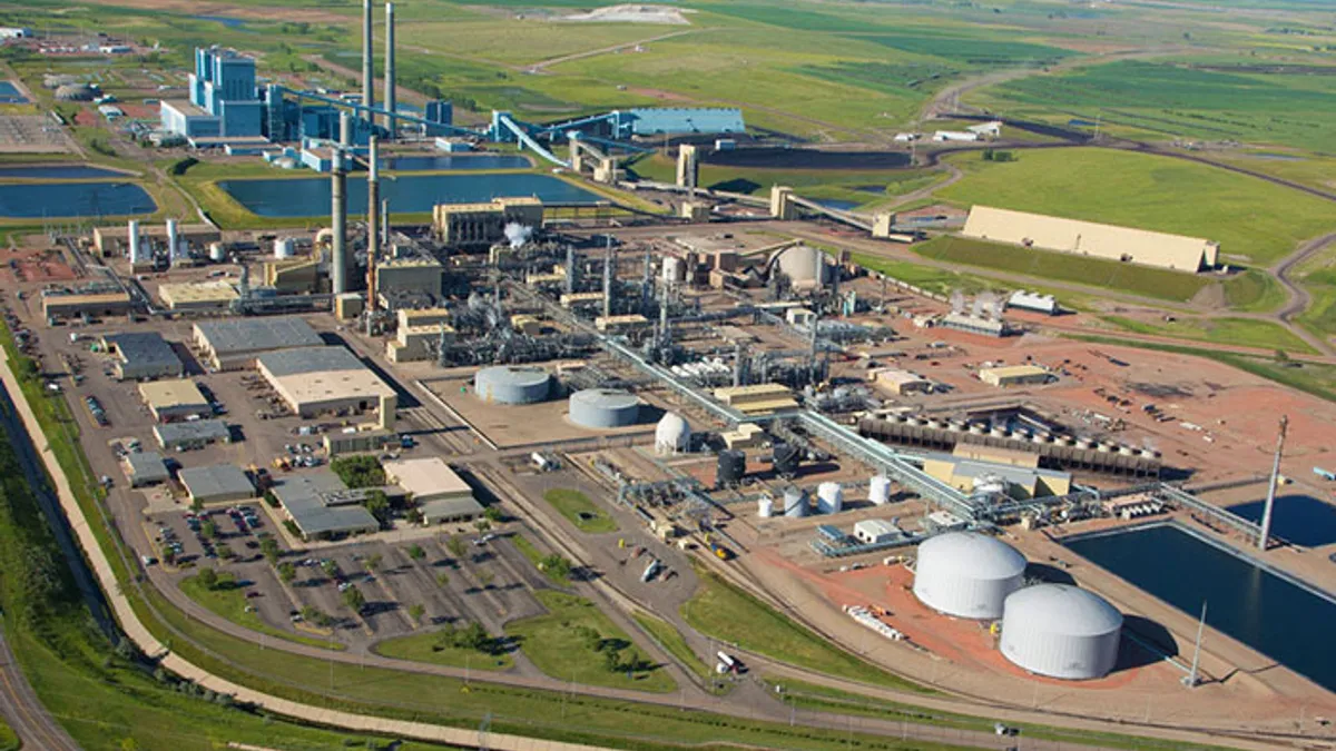 An aerial view of an industrial complex with buildings, storage tanks and pipes, and a power plant in the background.