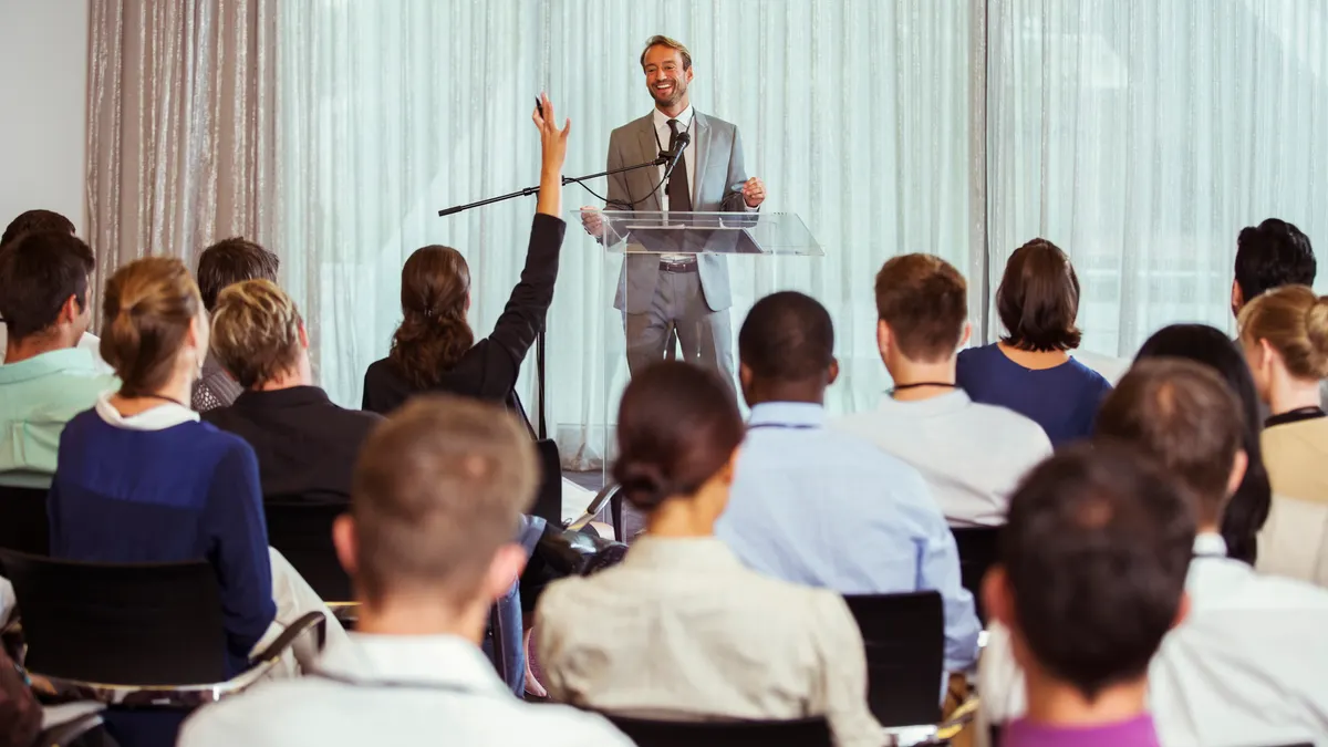 Businessman giving speech in conference room, woman from audience raising hand.