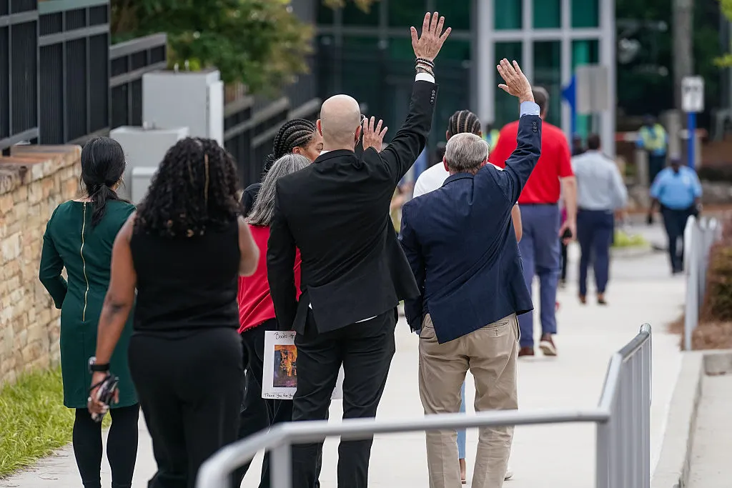 Two men in suits stand in the center of a group of people walking down a side walk with their right hands in the air while a woman in front one man on the left raises her right hand.