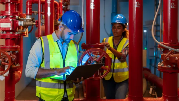 A building engineer uses his laptop to inspect the pipeline and valve of service water supply in a fire riser room, while a technician in the background tests a fire suppression system.