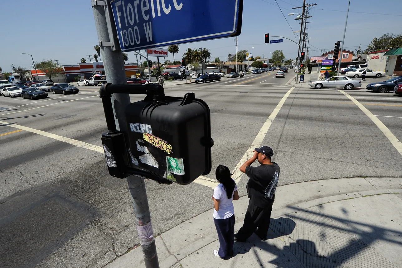 Two people wait on the curb before crossing a major intersection in Los Angeles.