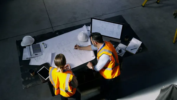 Two people in construction gear meet over a table with drawings and computers.