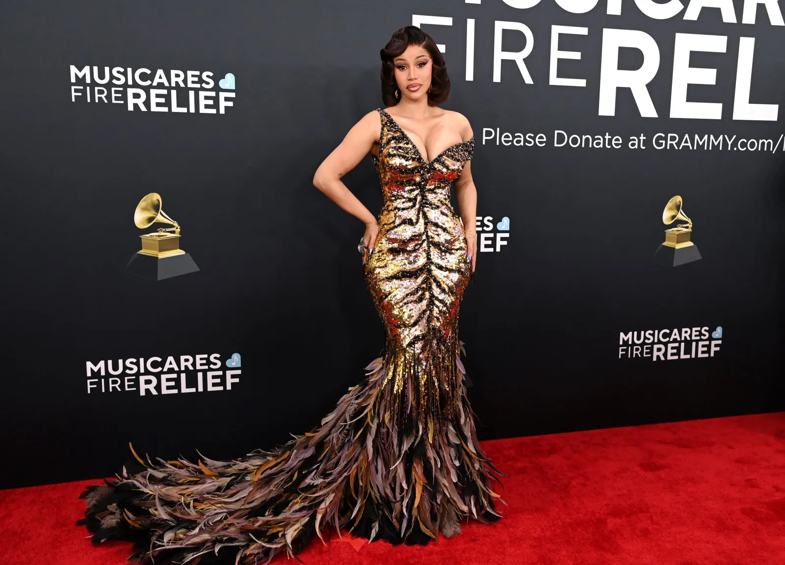 A person in a long sequined gown with a feather train stands on the red carpet.