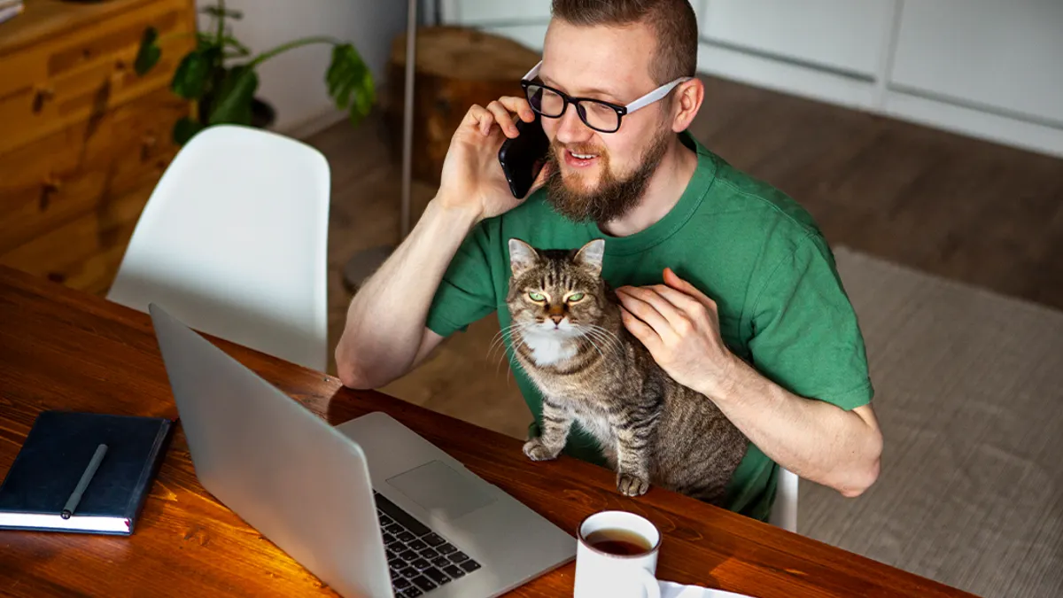 A man on his computer petting a cat and talking on the phone