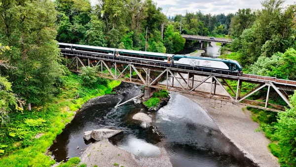 A green, white and brown train crosses a bridge over a river surrounded by green trees.