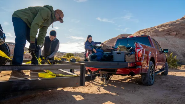 Several people shown securing cargo in a 2024 Ford F-150 pickup and preparing yellow hold-down straps on an attached trailer.
