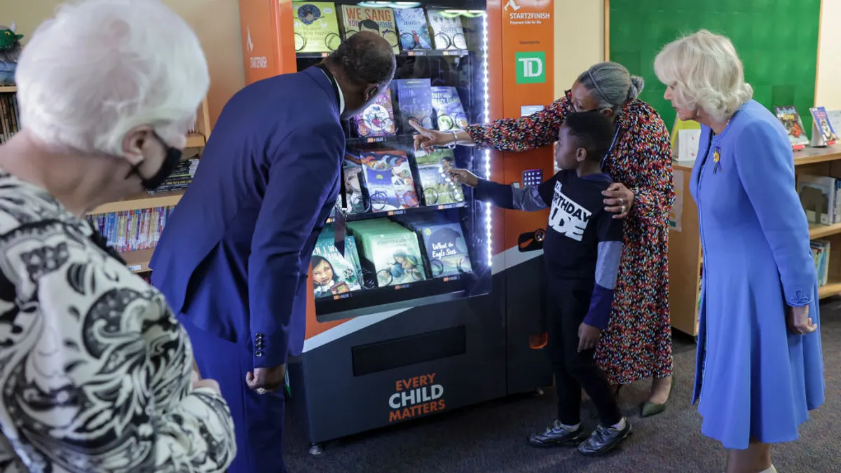 Four adults and a child stand in front of a book vending machine in a school as the child makes a selection.