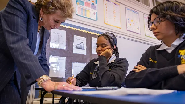 An adult is standing over a desk of a student in a classroom. The adult is looking at papers on the desk. Another student seated at another desk looks on.