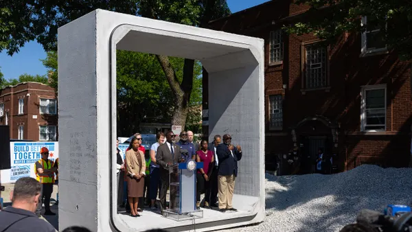 A person speaks at a podium surrounded by other people standing inside a massive concrete square.