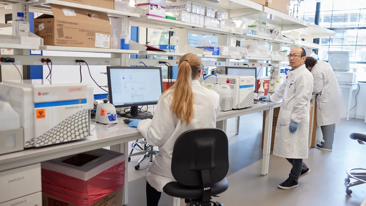 Two lab scientists work inside a research facility. A man turns to talk to his colleague, who is seated.