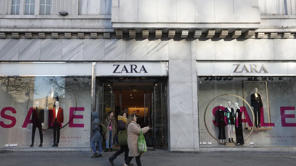 People walk past a Zara storefront, where the window displays are advertising a sale.