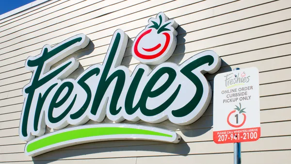 A photo of a logo on the exterior wall of a white building. The logo says Freshies. A sign is in the foreground that says Freshies online order curbside pickup only.