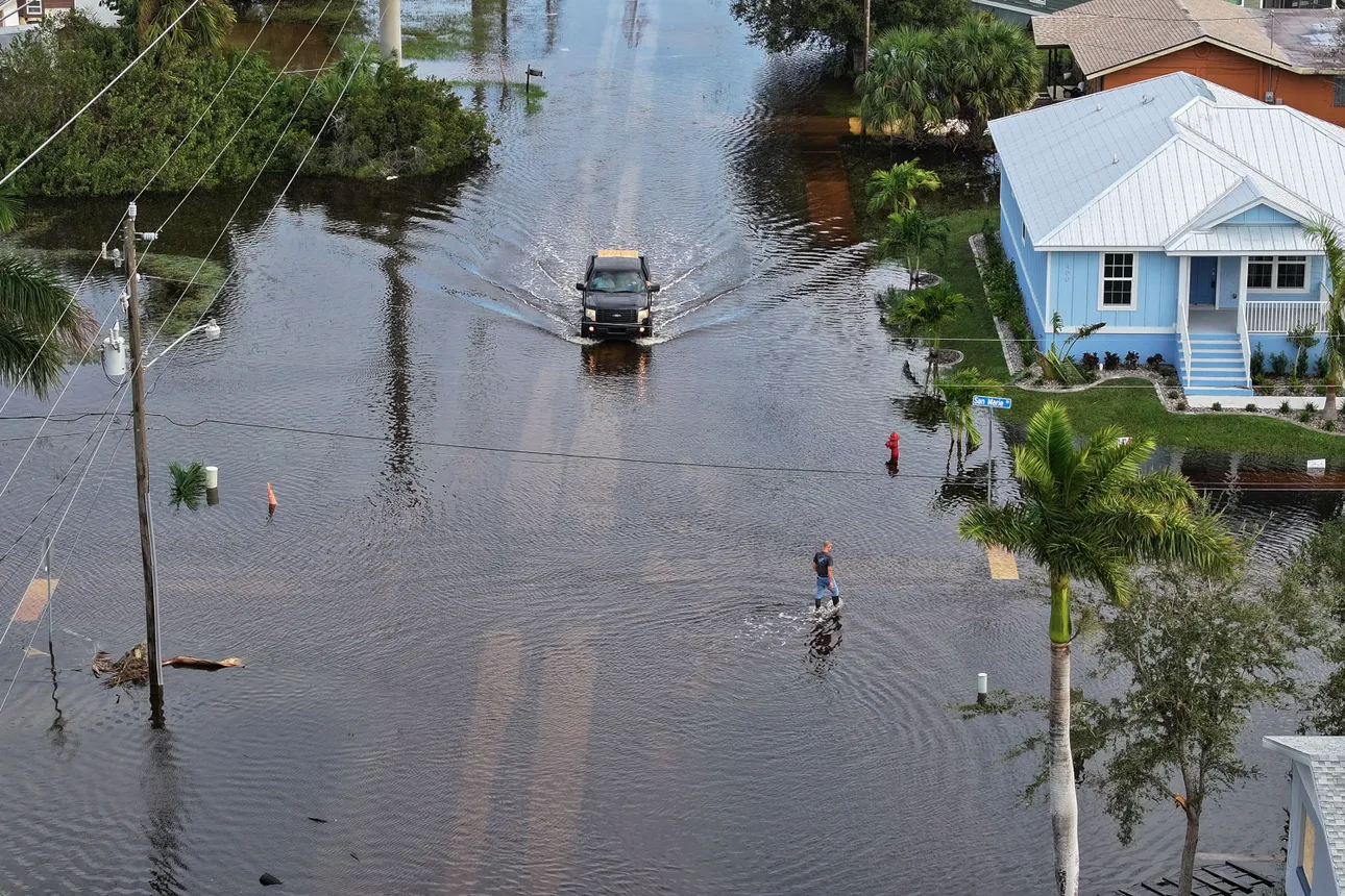 A vehicle drives in the middle of a flooded road, with a house, trees and a pedestrian around it.