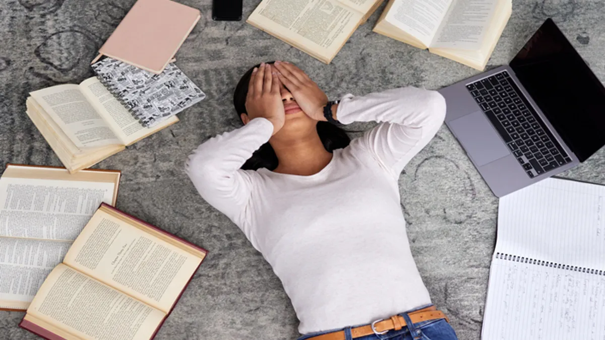 A student is laying on the floor with their hands over their face. A laptop, books, a phone and notebooks are on the floor surrounding the person.